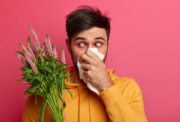 Frustrated man blows nose in tissue, has redness around eyes, symptoms of allergy, has unhealthy look, concentrated at blooming flower, suffers from rhinitis, allergic reaction. People and illness
